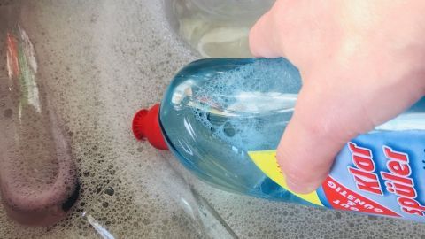 Eine Hand hält eine Flasche Glanzspüler über einem Spülbecken mit schaumigem Wasser und Gläsern im Inneren.