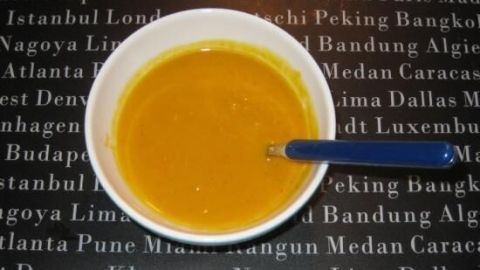 Eine scharfe Hokkaido-Kürbis-Suppe wird in einer weißen Schüssel serviert, daneben liegt ein blauer Löffel auf einem schwarzen Hintergrund mit Städtenamen.