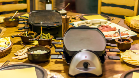 Ein Raclette-Grill steht in der Mitte eines festlich gedeckten Tisches, umgeben von Zutaten wie Würstchen, Käse und Salzgurken.
