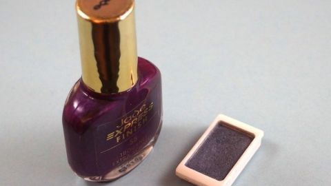 Ein Nagellack in kräftigem Lila steht neben einem alten Lidschatten in einem kleinen, rechteckigen Behälter auf einem neutralen Untergrund.