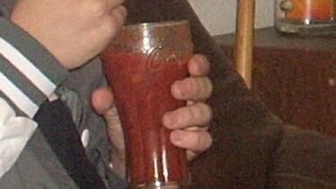 Ein Glas alkoholfreier Bloody Mary wird in einer Hand gehalten, während sich eine andere Hand mit einem Löffel nähert. Im Hintergrund ist ein Sofa sichtbar.