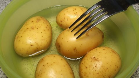 Fünf Kartoffeln liegen in einer grünen Schüssel mit Wasser, während eine Gabel in eine der Kartoffeln gestochen wird.