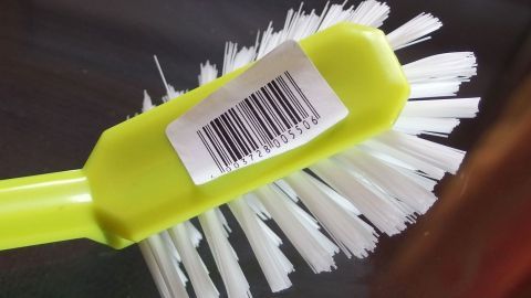 Ich habe keine Lust mehr dazu, erst alle möglichen Tricks zur Entfernung auszuprobieren. Wenn ich zum Beispiel ein paar Schuhe kaufen möchte, sage ich ganz freundlich: "Dieses Paar hätte ich gern. Aber bitte entfernen Sie doch den Aufkleber". Eine grüne Bürste mit weißen Borsten liegt auf einem Tisch, sichtbar mit einem lästigen Aufkleber und Barcode.