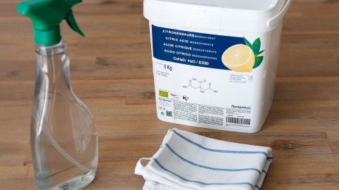 Sprühflasche und Tuch auf Holztisch neben 5 kg Zitronensäurebehälter, Text: "ZITRONENSÄUREMONOHYDRAT, CITRIC ACID MONOHYDRATE".