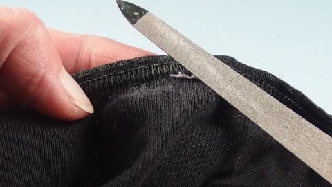 Eine Hand hält ein Kleidungsstück, während ein Werkzeug Piksende Nähte entfernt, im Kontext eines hellen Hintergrunds.