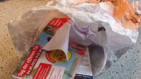 Eine zerrissene Bäckertüte liegt in einer Schüssel, während darin auch bunte Werbeflyer zu sehen sind, die Abfall zeigen.