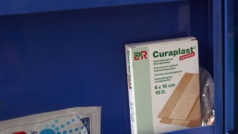 In einem blauen Schrank ist ein Paket Curaplast verbunden mit einem Verband sichtbar, während andere medizinische Utensilien daneben liegen.