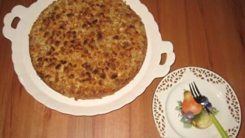 Ein Apfelkuchen mit goldbrauner Kruste liegt auf einem weißen Teller und wird neben einer Schale mit Obst serviert.