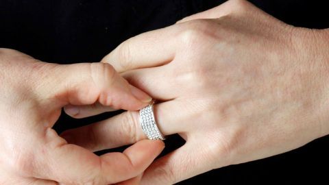 Mit lauwarmen Seifenwasser bekommst du deinen Ring abends wieder leichter vom Finger. Eine Hand zieht einen engen Ring von einem Finger ab, während eine andere Hand unterstützt, vor einem dunklen Hintergrund.