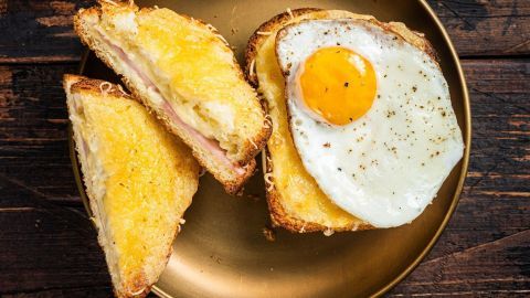 Ein golden gebratens Frühstücksei liegt auf geröstetem Brot, umgeben von geschmolzenem Käse und Schinken.