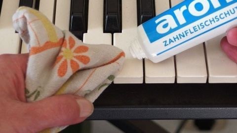 Eine Hand hält ein Tuch, das mit Zahnpasta benetzt wird, um Klaviertasten zu reinigen. Der Hintergrund zeigt Klaviertasten.
