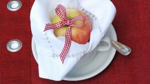 Kreiere eine einladende Tischdekoration mit einem dekorativen Besteck-Arrangement, wie im Artikel beschrieben. Die hübsch eingewickelte Apfel-Dekoration bringt einen rustikalen Charme in deine Kaffeetafel und ergänzt den versunkenen Apfelkuchen perfekt. Ein weißes Tuch mit einem Apfel, dekoriert mit einem roten Band, liegt auf einem Teller auf einem roten Tisch.
