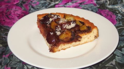 Ein Stück Obstkuchen liegt auf einem weißen Teller. Der Kuchen hat eine goldene Kruste und ist mit Obst belegt.
