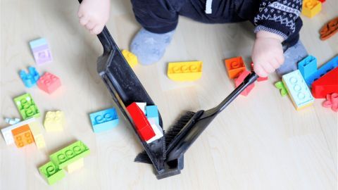 Ein Kind räumt fröhlich mit einem Besen bunte Lego-Bausteine auf, die verstreut auf einem hellen Fußboden liegen.