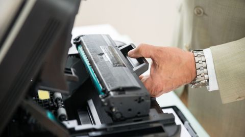 Ein Hand greift eine Tonerkartusche und setzt sie in einen Drucker ein, während die Umgebung hell und modern wirkt.