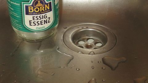 Essigessenz-Flasche steht neben einem Abfluss in einem nassen, metallischen Spülbecken. Text auf Flasche: "Born Essig Essenz Classic".