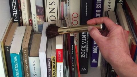 Ein Make-up-Pinsel wird verwendet, um Staub von Büchern in einem Regal zu wischen.