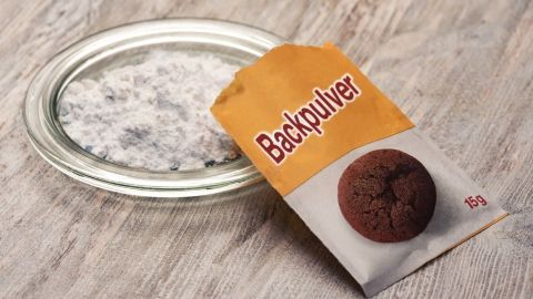 Ein Päckchen Backpulver liegt neben einer Schale mit weißem Pulver auf einer Holzoberfläche, bereit zur Anwendung gegen Warzen.