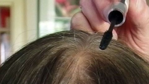 Eine Hand hält einen Applikator, der Wimpernfarbe auf den grauen Haaransatz aufträgt, um ihn zu decken.