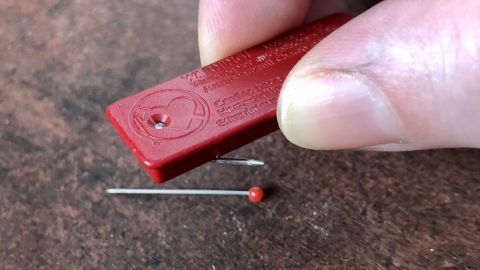 Wenn dir Stecknadeln oder Nadeln auf den Boden fallen, kannst du sie mit einem Magneten im Handumdrehen aufsammeln! Meistens hat man einen an der Kühlschranktür hängen. Eine Hand hält einen roten Magneten über eine Stecknadel, um diese von einer Oberfläche aufzuheben.