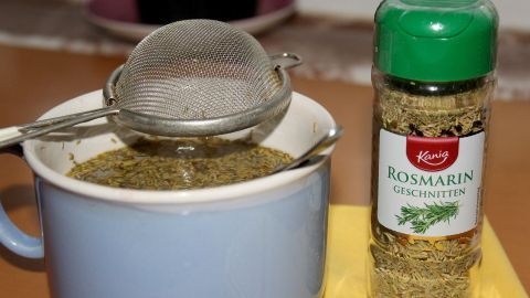Ein blauer Becher mit frisch aufgebrühtem Rosmarintee steht neben einer Gewürzflasche und einem Sieb zum Abseihen.