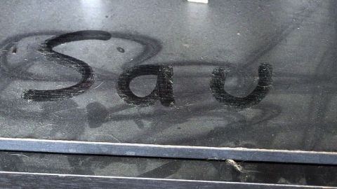 Auf einer dreckigen Oberfläche steht das Wort "Sau", während Staub und Dreck im Hintergrund sichtbar sind.