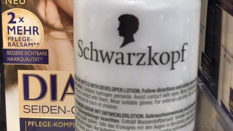 Eine Flasche Haarfärbemittel ist im Vordergrund sichtbar, während im Hintergrund eine Frauenansicht und Produktbeschreibung zu sehen sind.