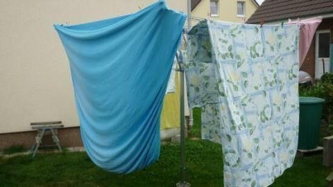 Ein blaues und ein gemustertes Spannbettlaken hängen an einer Wäschespinne im Garten, während es bewölkt ist.