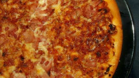 So kann man Pizza einpacken und transportieren, ohne dass der Belag an der Folie kleben bleibt. Eine frisch gebackene Pizza liegt auf einem schwarzen Teller, der Belag besteht aus Käse und Schinken, appetitlich und knusprig.