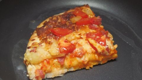 Eine Pizzastücke liegt in einer Teflon-Pfanne und wird erhitzt, um wieder knusprig zu werden, in einer einfachen Küche.