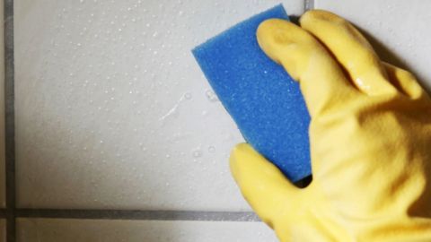Eine Hand mit einem gelben Handschuh reinigt helle Fliesenfugen mit einem blauen Schwamm in einem Badezimmer.