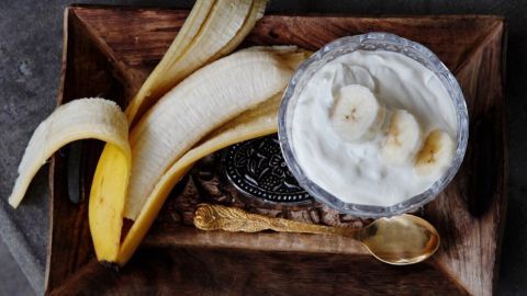 Ein Becher mit cremigem Joghurt und Bananenscheiben liegt neben einer halbgeschälten Banane auf einem rustikalen Tablett.