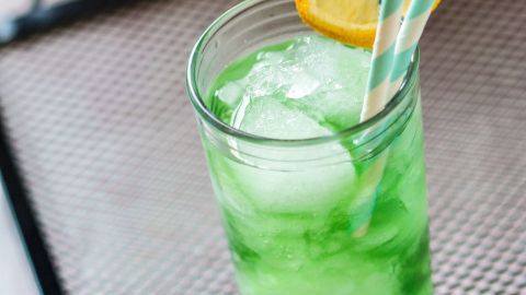 Ein erfrischendes Getränk in einem Glas, gefüllt mit Eis und grünem Sirup, steht auf einem Tablett im Außenbereich.