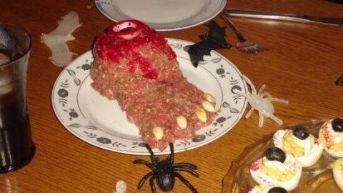 Eine blutige Fußform aus Hackfleisch liegt auf einem Teller, umgeben von Spinnen und anderen Halloween-Dekorationen.