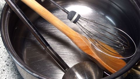 Ein Löffeltopf mit lauwarmem Wasser enthält Kochwerkzeuge wie einen Holzlöffel, einen Schneebesen und einen Schöpflöffel.