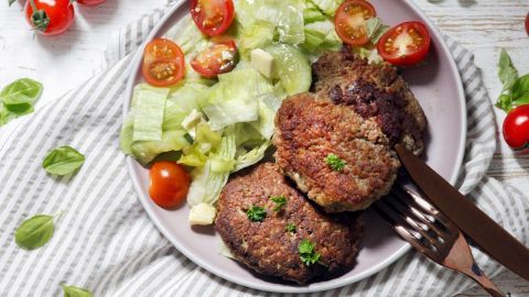Saftige Hackfleisch-Pattys liegen auf einem Teller, umgeben von frischem Salat und Tomaten, auf einem gestreiften Tuch.