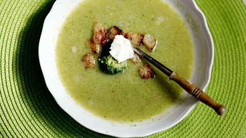 Verfeinere eine zu scharfe Suppe mit einem Schuss Öl, um die Schärfe zu mildern. Dieses Bild zeigt eine cremige Suppe mit Brokkoli und Croutons, perfekt abgerundet mit etwas Sahne obendrauf. Suppe mit Croutons und einem Klecks Sahne in weißer Schüssel, auf grünem Platzdeckchen, neben einem Holzlöffel.