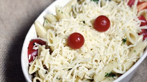Das Überstreuen direkt aus dem Beutel verleitet dazu, sehr üppig und verschwenderisch mit den leckeren Streuseln umzugehen. Da hilft es, den großen Beutel Fertigkäse von vornherein zu rationieren und die Hälfte für den nächsten Schlemmertag aufzuheben. Ein Auflauf mit Pasta, garniert mit Kirschtomaten und rationiertem Streukäse, steht auf einem rustikalen Tuch.