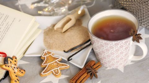 Eine Tasse Gewürztee steht auf einem dekorativen Tisch neben Keksen, Zucker und einer offenen Buchseite im Hintergrund.