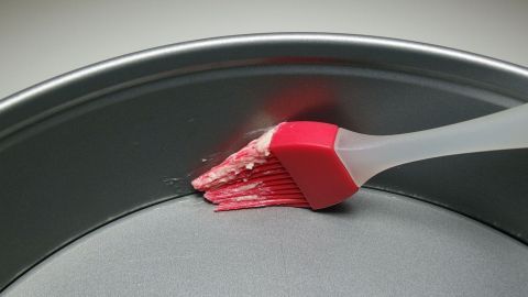 Ein Pinsel mit rotem Silikon fettiert gleichmäßig die Innenfläche einer runden Backform aus Metall, vor einem neutralen Hintergrund.