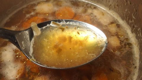 Ein Löffel voller Brühe hebt sich aus einem Topf und zeigt klare Flüssigkeit und Gemüsestücke in der heißen Suppe.