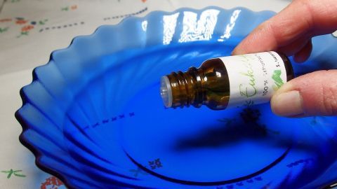 Eine Hand hält eine Flasche Eukalyptusöl über einer blauen Schale, bereit, es in heißes Wasser zu geben.