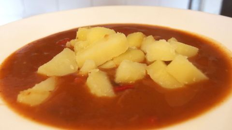 Eine Tomatensuppe mit Kartoffelstücken ist in einer weißen Schüssel angerichtet und vor einem neutralen Hintergrund platziert.