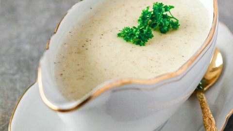 Die Béchamelsauce befindet sich in einem eleganten, goldrandigen Kännchen, garniert mit frischer Petersilie auf einem weißen Teller.