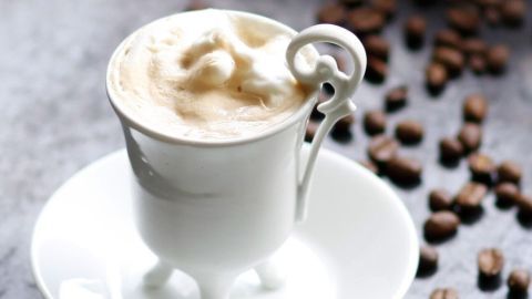Eine stilvolle Tasse mit cremigem Kaffee steht auf einem Teller, umgeben von Kaffeebohnen auf einer eleganten Oberfläche.