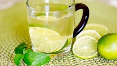 Ein transparentes Glas mit Pfefferminz-Apfel-Tee steht auf einem grünen Tisch, umgeben von Limettenscheiben und frischer Minze.