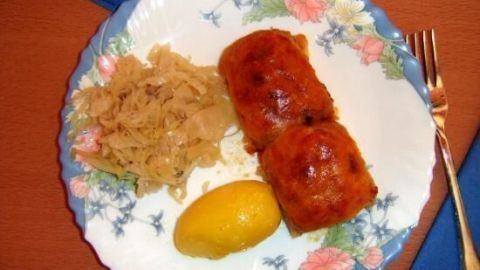 Zwei gebackene Speckknödel liegen auf einem floral dekorierten Teller neben Sauerkraut und einer Zitronenscheibe.