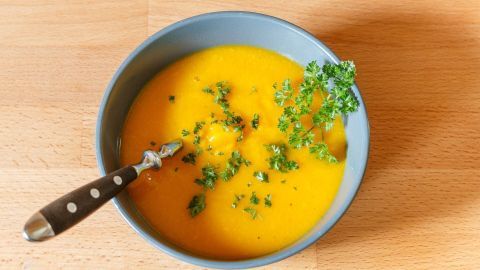 Eine Schüssel mit cremiger Kürbissuppe steht auf einem Tisch, garniert mit frischen Kräutern und einem Löffel.