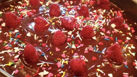 Eine schokoladige Sachertorte ohne Mehl ist dekoriert mit frischen Himbeeren und bunten Streuseln auf einer Tortenplatte.