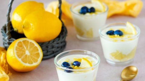 Kleine Gläser mit Mascarpone-Zitrus-Creme stehen auf einem Tisch, umgeben von Zitronen und einer Körbchen.